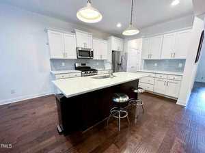 Modern kitchen with white cabinets, stainless steel appliances, a large island with a sink, and two metal stools. Pendant lights hang above the island, and the floor is dark wood.