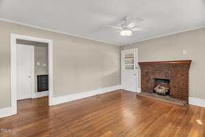 A living room with light beige walls, white trim, a ceiling fan with light, hardwood floors, a brick fireplace with a small stack of firewood, and a white door leading outside.