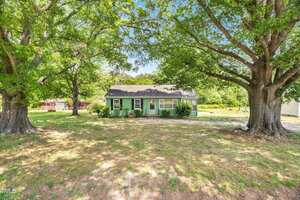 A single-story green house with a gray roof sits under large leafy trees at 2677 Nc-39 in Henderson, surrounded by a spacious yard with patchy grass and scattered shade. A red shed and open land are visible in the background.