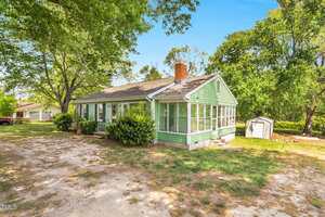 A small, single-story green house with white trim and a sunroom sits on a patchy lawn with trees and shrubs; a white shed stands in the background under a clear blue sky.