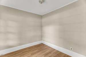 An empty room with beige walls, white trim, a ceiling light fixture, wood flooring, and an electrical outlet near the floor. No furniture or decorations are present.
