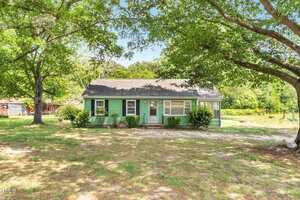 A single-story green house with white trim sits under large leafy trees. The front yard has patchy grass and a concrete walkway leading to the door. Shrubs are planted near the house, and sunlight filters through the trees.