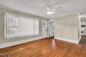 A bright living room with hardwood floors, beige walls, white trim, a ceiling fan with light, large vertical blinds covering a window, and a door with glass panels. The kitchen is visible in the background.