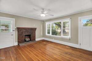 A living room with hardwood floors, a brick fireplace, beige walls, a ceiling fan, vertical blinds on a large window, and a white door leading outside. Natural light fills the space.