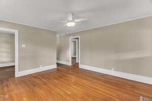 A room with beige walls, white trim, and a wooden floor. There is a ceiling fan with a light, and three doorways leading to other rooms. The space is empty and well-lit by natural light.