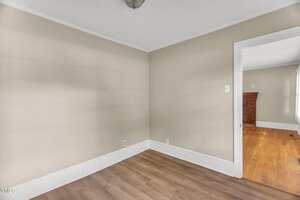 Empty room with beige walls, white trim, and light wood flooring. There is a doorway leading to another room with hardwood floors, a brick fireplace, and a window with white curtains. Ceiling light fixture is visible.
