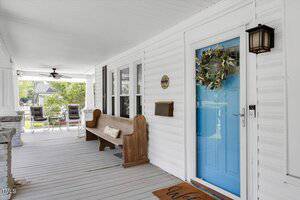 A front porch with white siding, a bright blue door decorated with a wreath, a wood bench with a pillow, a brown welcome mat, and outdoor chairs and a ceiling fan visible in the background.