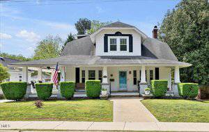 A white, one-and-a-half-story house with black shutters on 310 Henderson Street, Oxford. It features a covered front porch, dormer window, American flag, and two trimmed hedges flanking the walkway to the front door.
