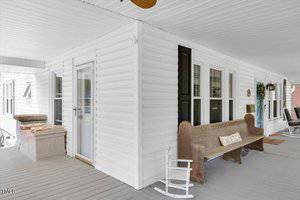 A white house porch features a wooden bench with a decorative pillow, a small white rocking chair, and a storage chest. The porch is covered, with white siding and gray flooring.
