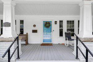 A white house front porch with black railings, a light blue door with a wreath, a welcome mat, white rocking chair, mailbox, house number 310, and several windows with white blinds.