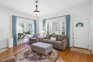 A living room with two gray sofas, a matching ottoman, a round rug, wood floors, and blue curtains on windows. There is a white front door, a floor lamp, and potted plants near the windows.