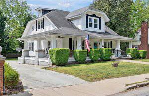 A white two-story house with a large wraparound porch, black roof, and dormer windows, featuring an American flag on the porch and trimmed green hedges in the front yard. Concrete driveway and sidewalk are visible.