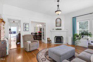 A living room with hardwood floors features a gray ottoman, a beige recliner, a gray sofa, a white brick fireplace, a round rug, and a window with teal curtains. A chandelier is visible in the adjacent room.