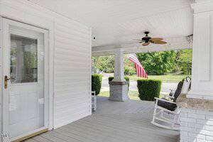 A white front porch with a ceiling fan, two rocking chairs, a white door with a screen, and an American flag in the yard. The porch has light gray flooring and white railings with brick columns.