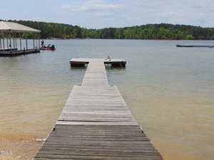 A wooden dock extends into a calm lake with a wooded shoreline in the background. A covered dock with a boat and two people is visible to the left, and another dock is seen in the distance to the right.
