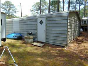 A large metal storage shed with a white door and diamond-shaped window sits on a pine-covered lot. A blue kayak, black grill, and concrete blocks are placed near the shed. Tall trees surround the area.