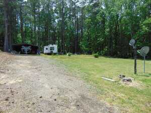 A gravel driveway leads to a small trailer and covered outdoor seating at 198 S Piney Grove Road, Manson. Set in a grassy clearing surrounded by trees, the yard also features two satellite dishes mounted on wooden posts.