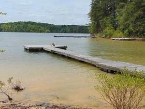 A wooden dock extends over a calm lake with cloudy water, surrounded by trees and vegetation. Another dock is visible in the background, and the sky is partly cloudy.