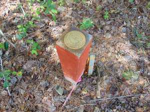 A rectangular red concrete survey marker stands in a wooded area with pine needles and leaves on the ground; a circular brass plaque with text is affixed to the top. A small orange and white post is next to it.