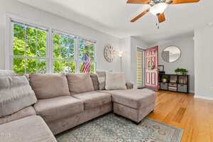 A living room with a beige sectional sofa, large windows showing greenery outside, a wooden floor, a ceiling fan with a light, a pink front door, and a console table with a round mirror and decor items.