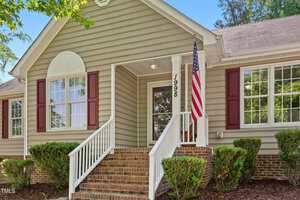 Single-story house with beige siding, brick foundation, red shutters, and white trim. Brick steps with a white railing lead to a covered front porch. An American flag is mounted near the entryway. Bushes border the walkway.