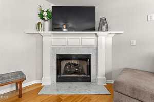 A white fireplace with a marble surround and hearth, a black fireplace insert, and a flat-screen TV on the mantel. Decorative items include a flower arrangement, a small potted plant, and a black wire-style vase.