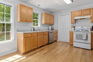 A kitchen with light wood cabinets, a white stove and hood, a dishwasher, and a double sink under a window; natural light enters through the windows, and the floor is light wood.