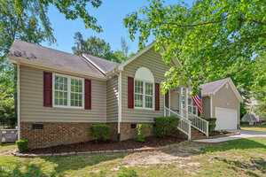 A single-story house at 1998 Ferbow Street in Creedmoor features beige siding, white trim, and maroon shutters. It has a brick foundation, manicured bushes, an American flag by the front steps, and an attached garage amid lush grass and trees.
