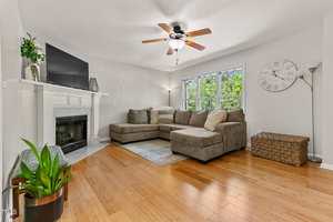 A living room with a gray sectional sofa, a TV on a white fireplace mantel, a ceiling fan, a floor lamp, a large wall clock, a woven basket, a potted plant, and wood flooring under a window with trees outside.