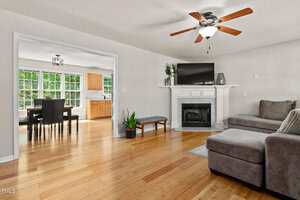 A living room with light wood floors, a gray sectional sofa, a white fireplace with a TV on top, a ceiling fan, a potted plant, and an adjoining dining area with large windows in the background.