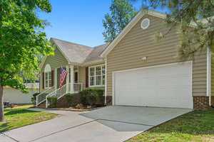 A beige single-story house with white trim, a two-car garage, a front porch with railing, an American flag, and a sloped driveway. There are bushes along the porch and trees around the yard.