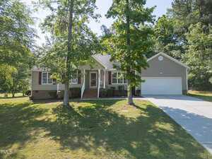 Single-story house with beige siding, white trim, and a two-car garage, partially shaded by tall trees in the front yard. An American flag hangs by the front porch. Concrete driveway leads to the garage.
