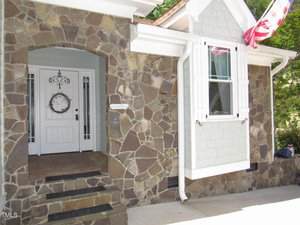 Front entry of a house with a stone facade, white door with decorative metalwork, a wreath, small steps, and a window with white shutters. Part of an American flag is visible at the top right.