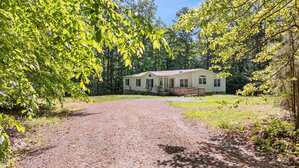 A single-story manufactured home with a small front porch sits at the end of a gravel driveway on 2690 Rivers Edge Road in Oxford, surrounded by trees and greenery on a sunny day.