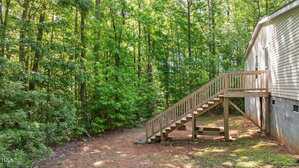 A wooden staircase leads from the side entrance of a house down to a dirt yard bordered by a dense forest of green trees. The house has light-colored siding and a block foundation.