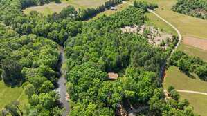 Aerial view of a rural landscape featuring dense green forest, a winding river, a few open fields, and a dirt road with a house partially visible among the trees.