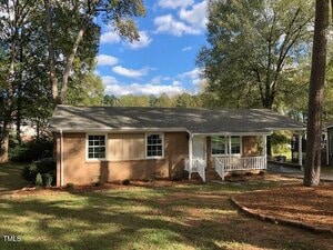 Charming single-story brick house at 157 Thorpe Street, Henderson, with a gray shingle roof, front porch with white railing, and large front window. Surrounded by tall trees and grass, driveway on the right, beneath a partly cloudy sky.