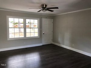 A room with light gray walls, dark wood flooring, a white door, a large window letting in natural light, and a ceiling fan with a light fixture. The room is empty and clean.
