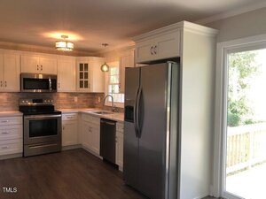 A modern kitchen with white cabinets, stainless steel appliances, wood-look flooring, tile backsplash, and a window above the sink. A glass door on the right leads to an outdoor deck with a view of trees.