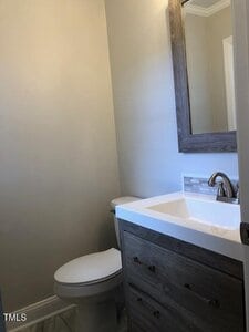 A small bathroom with a white toilet, a modern wood vanity with a white countertop, a rectangular mirror above the sink, and light-colored walls. There is a silver faucet and a tiled backsplash.