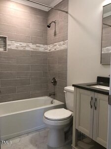 Modern bathroom with a bathtub-shower combo, gray tiled walls with a decorative accent strip, white toilet, and a vanity with a black countertop and mirrored cabinet above.