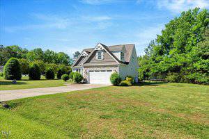 A two-story suburban house with white siding, a two-car garage, and dormer windows sits on a large, well-maintained lawn with trees and bushes under a clear blue sky.