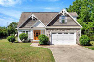 A two-story suburban house at 2062 Montgomery Drive, Franklinton features a stone and siding exterior, white garage door, wooden double front doors, triangular roof peaks, manicured bushes, and a concrete driveway on a sunny day.