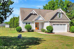 A two-story suburban house with beige siding, dark gable accents, and a double garage. The front yard is grassy with trimmed bushes, a rock, and a concrete driveway. Trees and a fenced area are visible in the background.