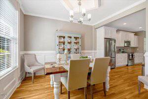 A dining area with a wooden table set for four, beige upholstered chairs, a glass-fronted china cabinet, and an adjoining modern kitchen with stainless steel appliances and white cabinetry. Light streams in from a window.