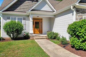 A concrete walkway leads to a wooden front door with glass panels on a light gray house with white trim, surrounded by green bushes and mulched flower beds.