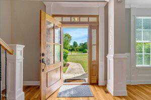 Open front door in a bright entryway, showing a welcome mat on hardwood floors and a view of a green lawn and trees outside under a blue sky. White trim and walls frame the doorway, and sunlight streams in.