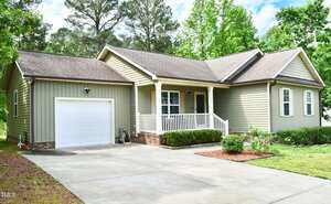Single-story house with light green siding, white trim, a covered front porch, and attached single-car garage. The front yard features bushes, a small garden bed, and a concrete driveway. Tall trees are in the background.