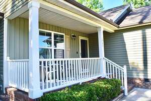 A small house porch with white railings, two chairs, and a table. The exterior walls are green with white trim, and the porch is supported by white columns. Brick steps lead up to the porch.