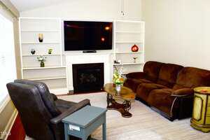 A living room with a brown sectional sofa, a dark armchair, a wooden coffee table, a side table, built-in white bookshelves, a flat-screen TV above a fireplace, and various decorative items on the shelves.